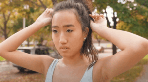 An advertisement depicting of a close up of a women in a blue tank top adjusting her ponytail. She is slightly sweaty and it is assumed she is jogging down the street. The streets are lined with trees. She looks straight ahead and looks determined.