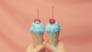 An advertisement of two hands holding two waffle cones containing scoops of blue ice cream with cherries and sprinkles on top. The backdrop is a peach colored curtain. The ice cream is dripping down the hands.