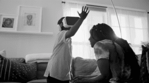 A black and white photograph of a woman and her child playing and laughing in their living room. The woman is facing away from the camera, looking up to her child. The child is reaching their arms up and wearing a Virtual Reality headset.