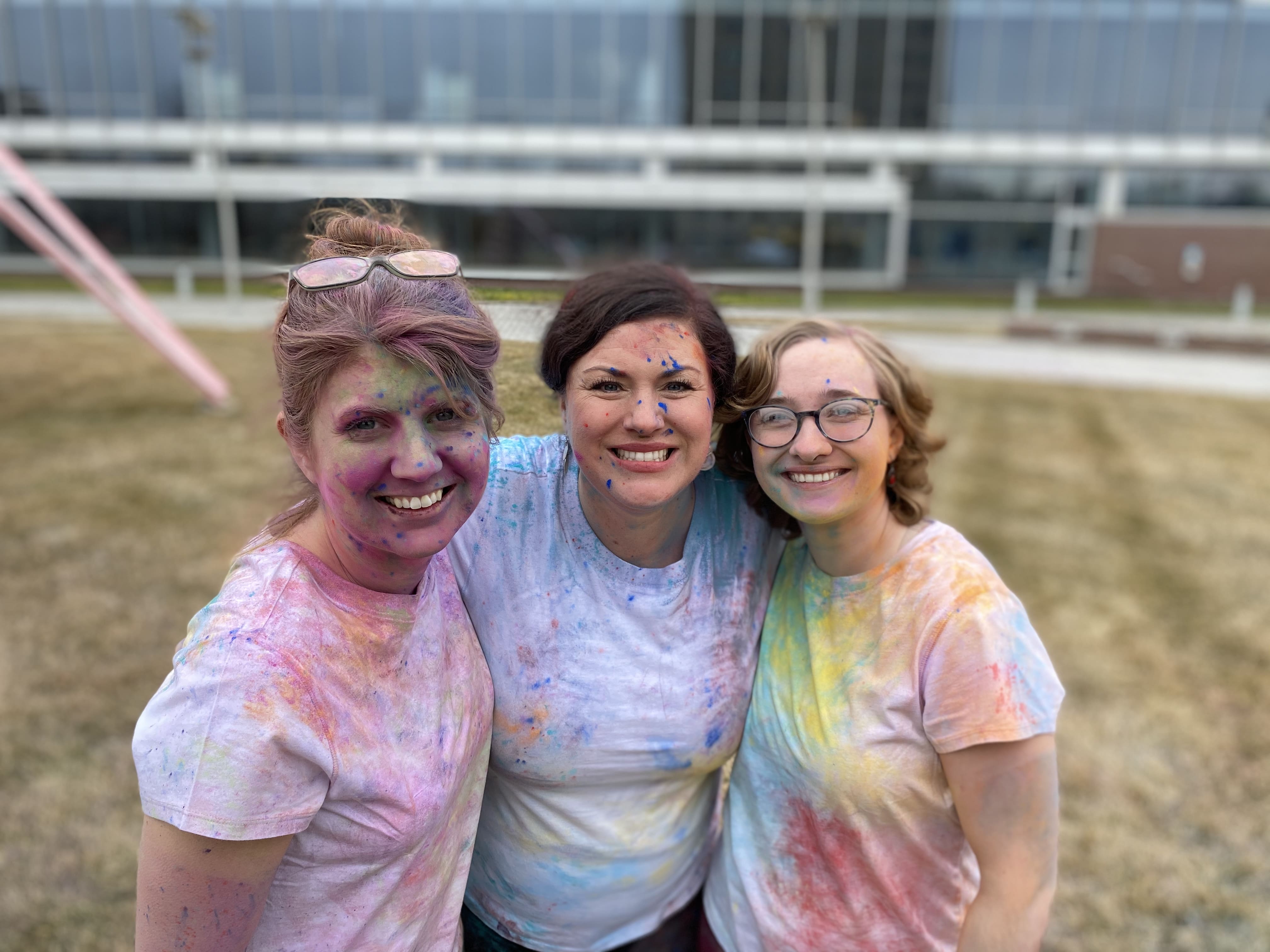 three students covered in colored paint holding each other outdoors
