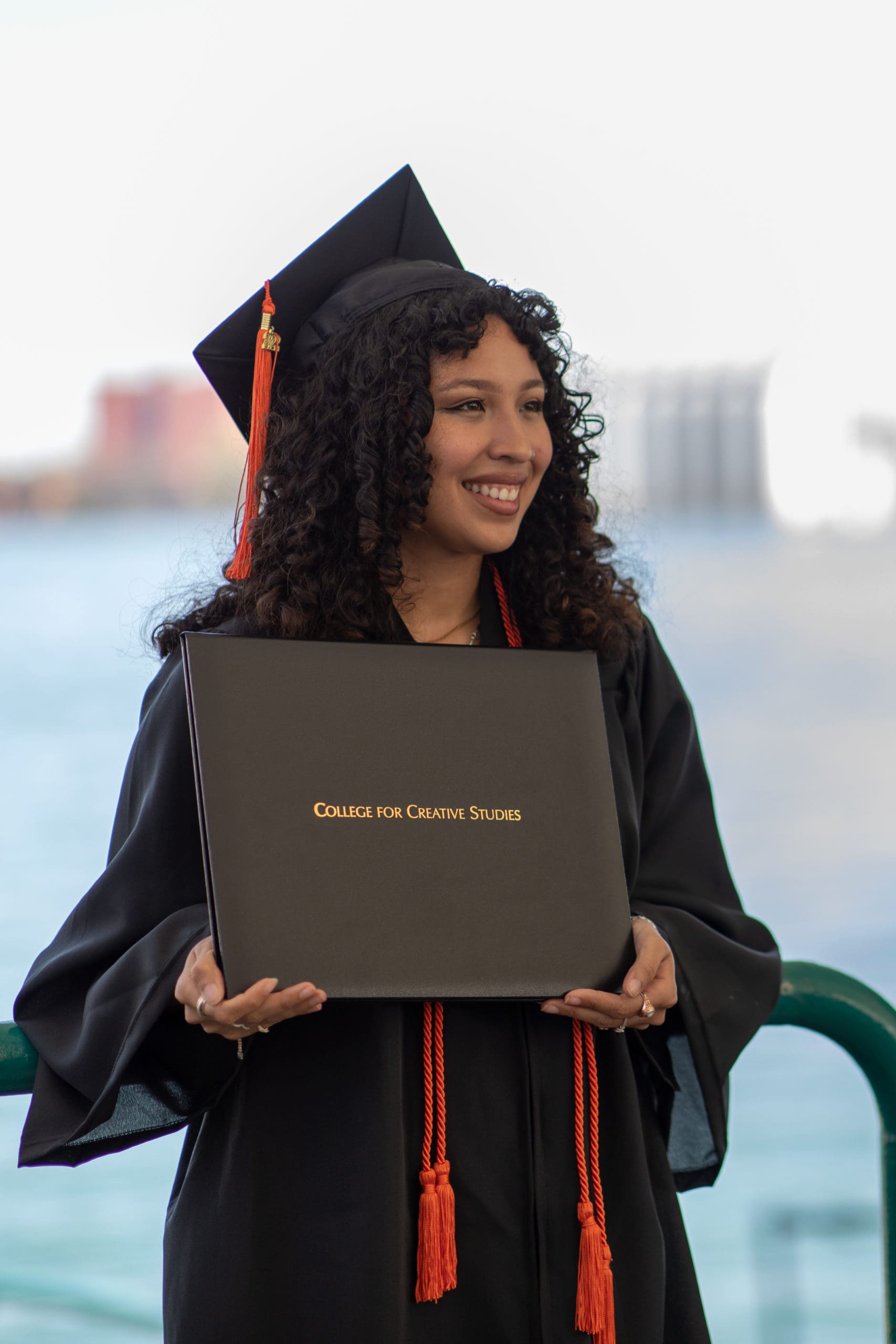 A graduating student with orange cords smiles for a photo and poses with their diploma.