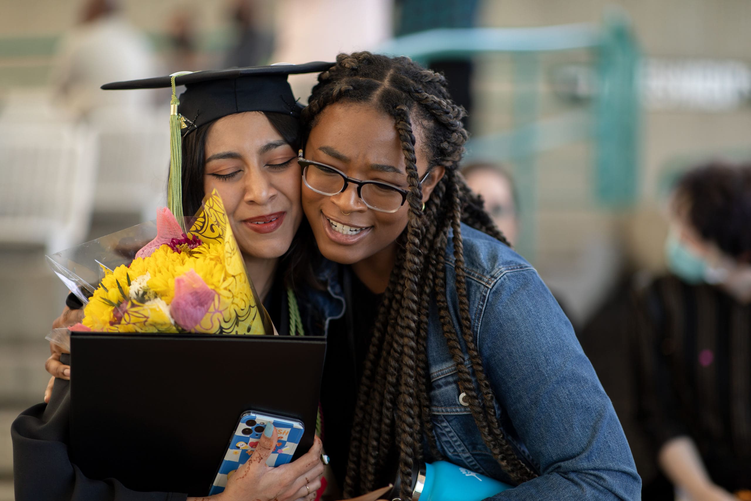 A graduating student hugs a loved one while holding their diploma and a bouquet of yellow and pink flowers.