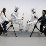 humans sitting alongside robotic figures, powering them with bicycles