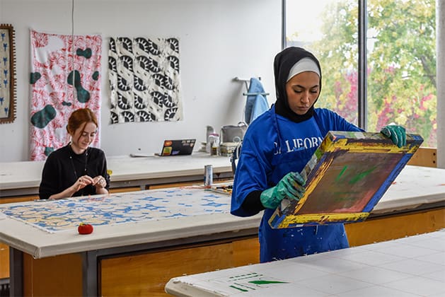 Students creating prints on fiber. One student holds a screen the other has her fabric lying on the table in front of her.