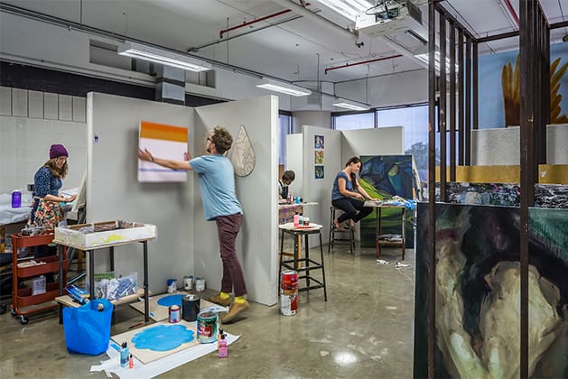 students hang artwork on modular walls, surrounded by paint and materials in a studio art and craft classroom