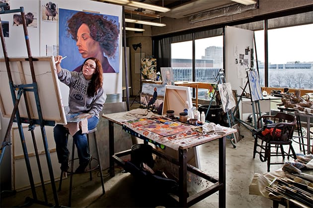 A student works on a large painting, surrounded by paints, materials and other large paintings in the painting studio