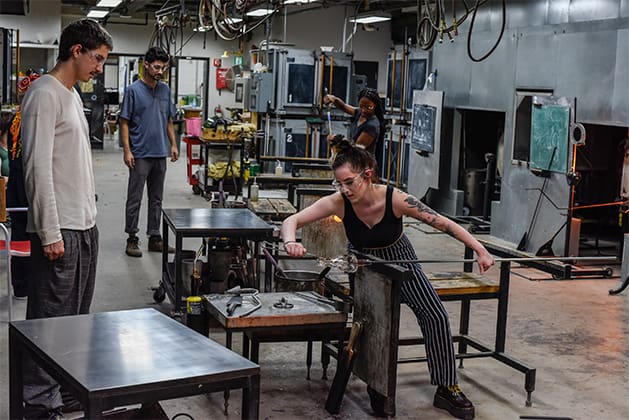 A student manipulates hot glass with metal tools while other students look on.