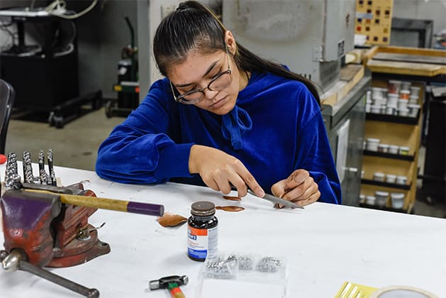 A student works on small metals and jewelry design with a file, surrounded by other metals tools on the table