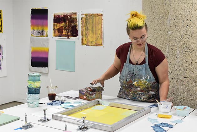 A student stands in front of a printmaking screen with yellow paint and other prints hung on the wall behind her