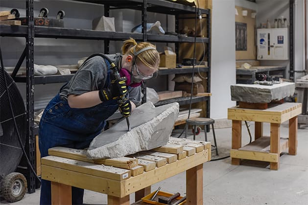 A student uses a tool to sculpt stone, surrounded by other sculptures and tools