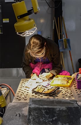 A student uses a large overhead light to work on a small-scale sculpture on a table with holes in it.
