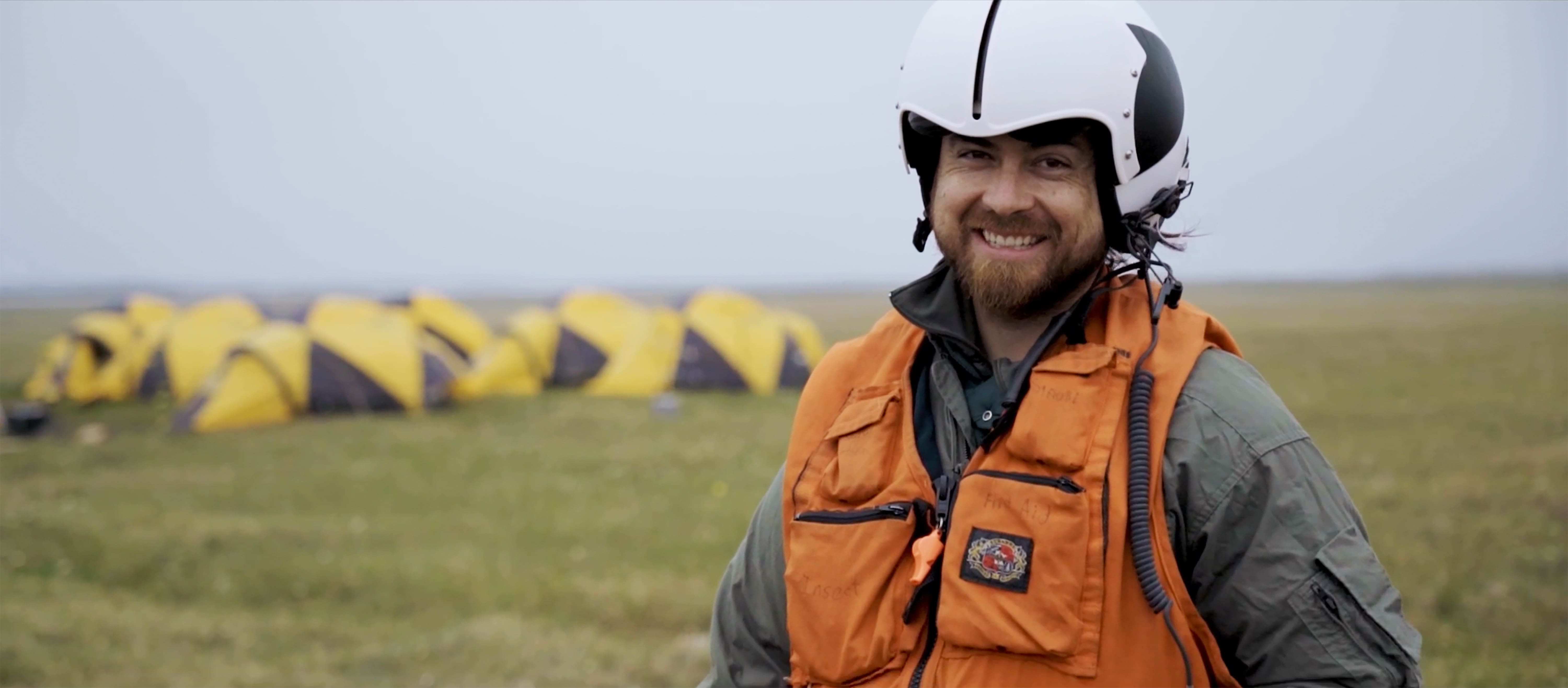 Artist Francis Vallejo is standing in an orange vest with a white helmet on his head in front of group of yellow tents in Alaska.
