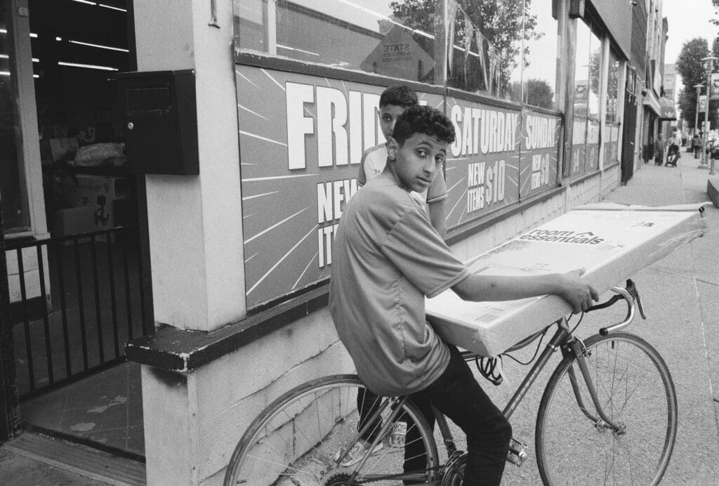 Black and white photo of two boys with a bike and a box leaving a store on the sidewalk