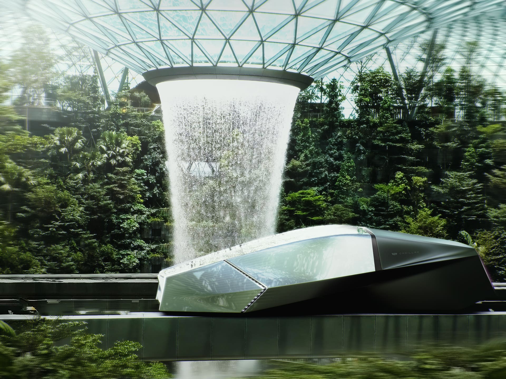 Concept art of a sleek silver and black vehicle driving on a track underneath a fountain waterfall. Located inside of a green house full of forestry