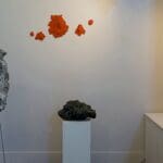 exhibition room with white walls. On the walls are orange and white installations pieces made of rough material. On stands are two more pieces made of rough grey material.