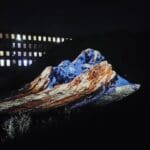 Photo of exhibition piece. An image of a blue, orange and white scene is projected onto a small hill in the nighttime.