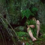 Photograph of a woman laying against a wall of mossy rocks, covering herself with grass.