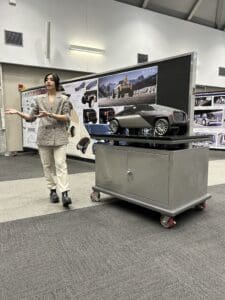 A student is standing in front of a large rectangular display board and a small table with a vehicle model.
