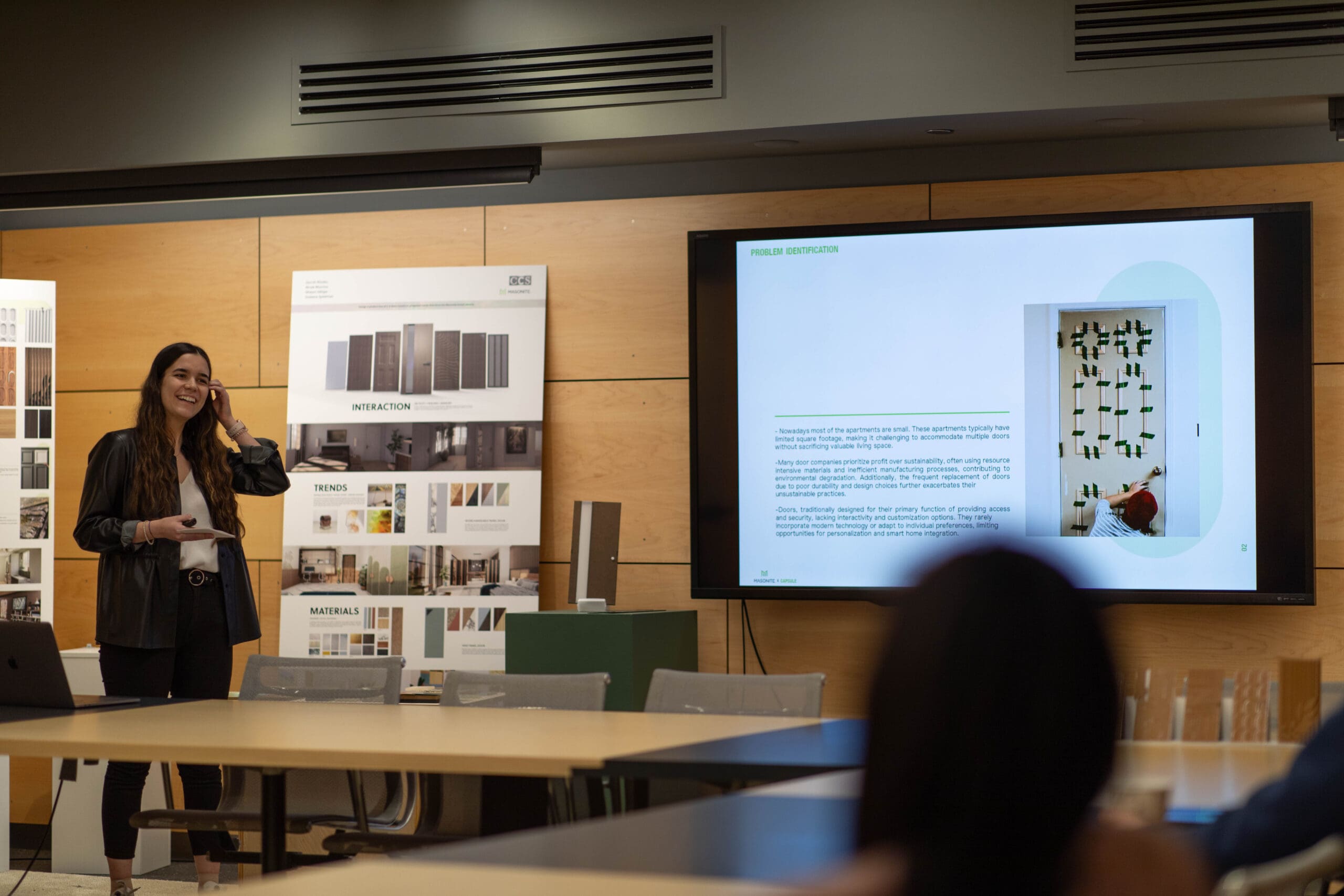 A student is standing in the front of the board room with their designs displayed on a large television. She is explaining her designs to a panel of seated judges.
