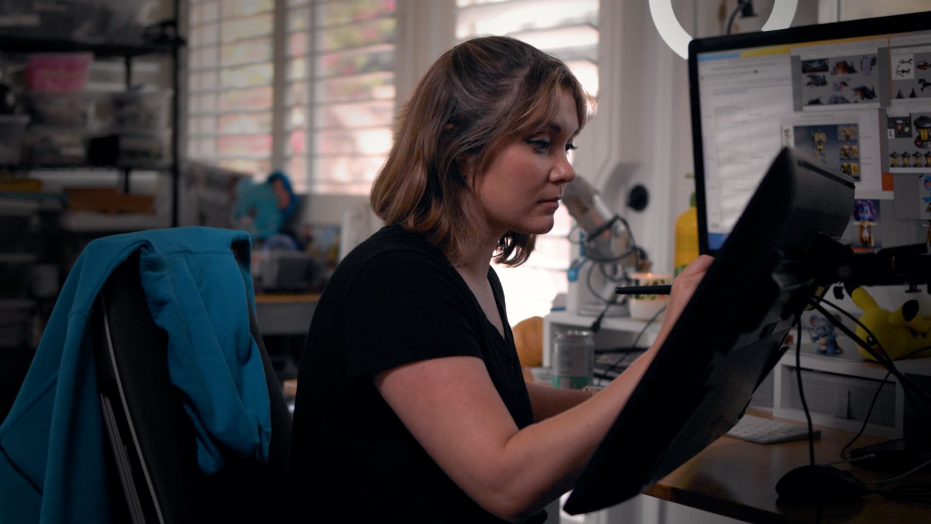 A woman is sitting in front of a computer and drawing with a stylus. She has a blue sweatshirt hung on the back of an office chair she is sitting on.