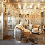 A room with mirrored walls, doors and cabinets that have a gold or metallic finish. There are two unique chairs in the center of the room, one covered with a fur throw, and a small table between them. A brown purse and pair of high heeled boots sit on the ground.