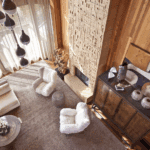 A view from above of a cozy living room with a large, textured fireplace. The room features a cream modular sofa, two fuzzy white armchairs, and wood-paneled walls.