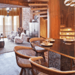 A cozy, wood-paneled dining and living area. The room features a glass dining table with unique, wire-frame chairs, a spiral staircase, and a view into a living space with two white, fluffy armchairs and a textured fireplace.