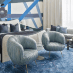 A bedroom with an abstract, blue and white painted accent wall behind a bed. The room has a large, blue rug, two pleated blue chairs, a grey couch, and large windows with white curtains.