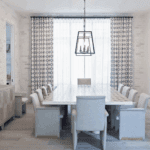 A bright dining room with a long table surrounded by upholstered chairs. The room has light wood floors, a light brick accent wall, and patterned curtains. A modern chandelier hangs over the table.