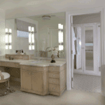 A bright bathroom with a double vanity and light wood cabinets. A large mirror spans the wall, and the counter is made of marble. A dressing room with mirrored doors is visible through an open doorway.