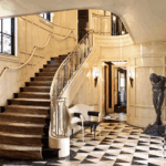 A grand staircase in an elegant entryway. The staircase has a curved metal railing and is lined with a runner. The floor has a black and white geometric pattern, and the walls are paneled in a glossy white. A large statue stands near the base of the stairs.