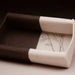 An art piece shaped like a shallow, open bowl. One half is dark brown and the other half is white with a delicate, black line drawing of bare tree branches.
