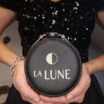 A person wearing a black sequined dress holds a small, round, black leather jewelry case with the gold logo "LA LUNE" and a half-moon symbol on it.