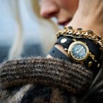 A close-up of a person's wrist wearing a layered watch and bracelet. The watch has a black face with a black leather strap, and a chunky gold chain bracelet is worn next to it.