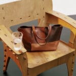 A close-up of a brown leather tote bag sitting on a wooden chair with a glass of water on the armrest.