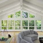 A sunroom with a vaulted ceiling and a large wall of windows looking out onto a lush forest. The room features a curved gray sofa, a telescope, and a toy truck and tractor on a rug. A small black dog sits near the rug.