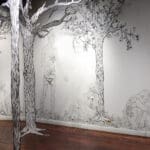 Four black and white figures covered in foliage are pictured, three kneeling on the ground, the other standing. They are black and white. Black and white trees are in the foreground, matching the trees on the wall with the figures.