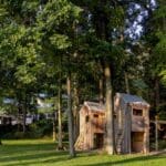 A two-tower wooden play structure connected by a swing set with a slide and a rope ladder, built on a grassy lawn surrounded by tall trees.