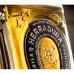 A close-up of a gold-colored bottle of Herradura tequila, focusing on the embossed label and the bottle's reflective surface.