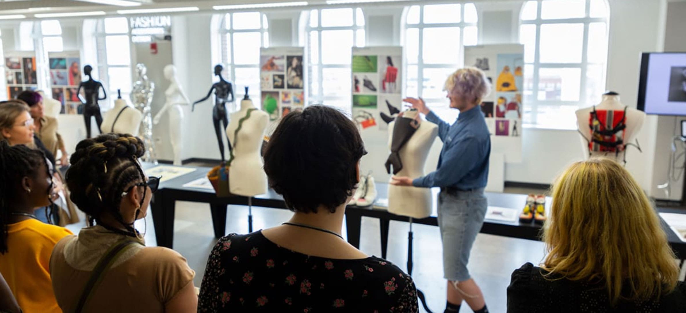 A group of CCS students are gathered around a long table that has fashion designs and mannequins displayed.