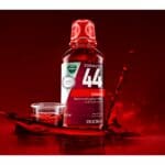 A Vicks Formula 44 cough syrup bottle sits in a puddle of red liquid, with a small cup of the liquid next to it. Liquid is splashing and dripping around the bottle and cup, as if the bottle is making a splash.