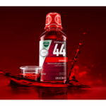 A Vicks Formula 44 cough syrup bottle sits in a puddle of red liquid, with a small cup of the liquid next to it. Liquid is splashing and dripping around the bottle and cup, as if the bottle is making a splash.