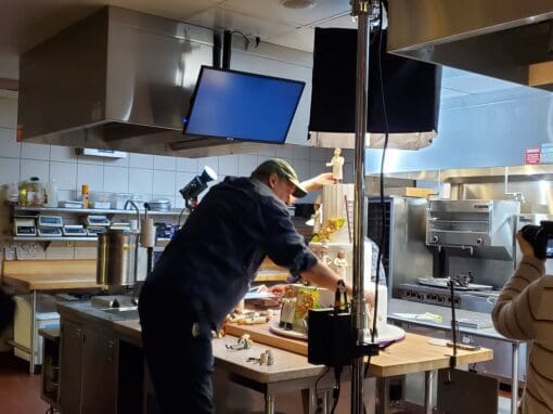 Adjunct Instructor Kevin Coyle working in a professional kitchen setting. He is standing at a counter, carefully arranging puppets and small objects for an animation shoot. A camera crew and professional lighting are visible on either side of him.