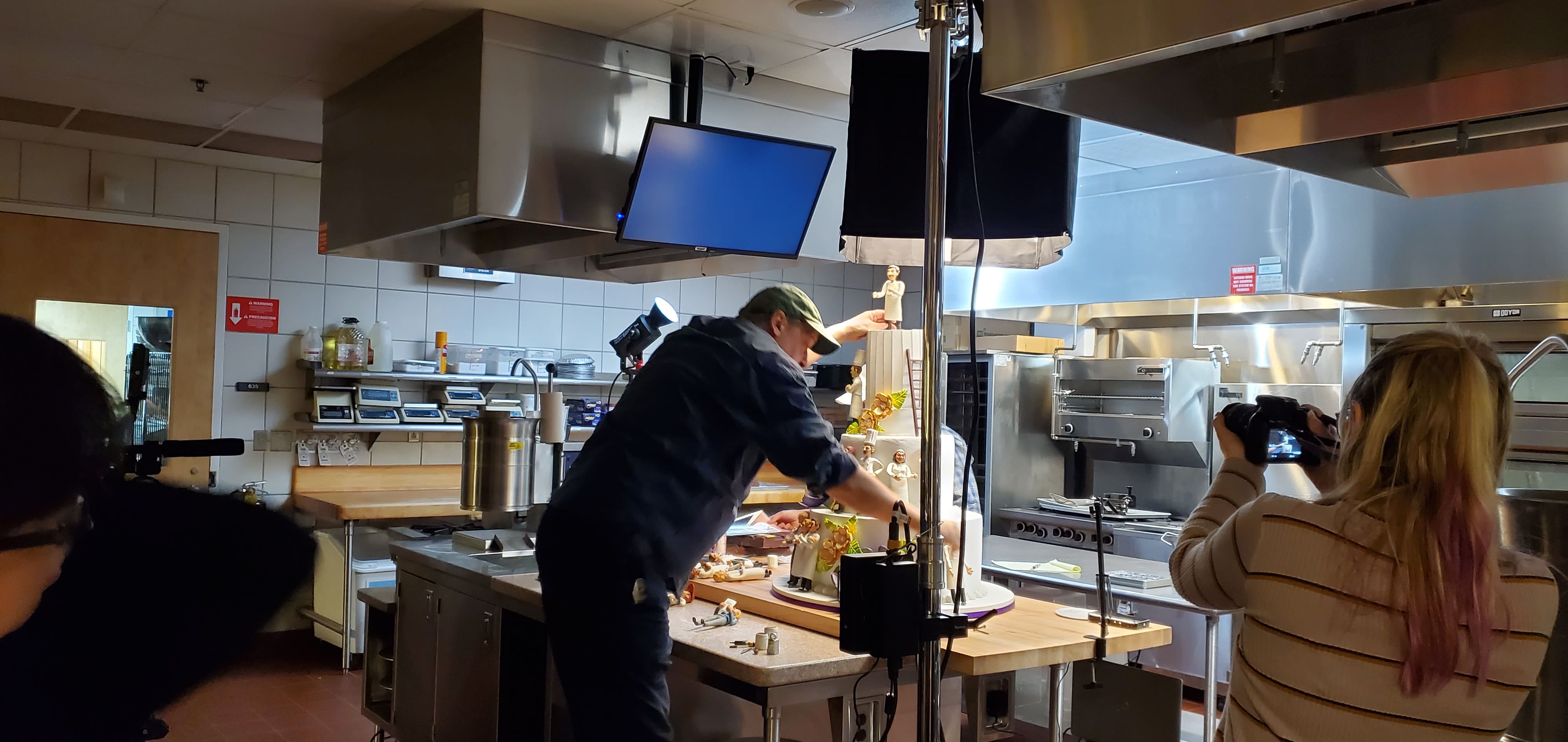 Emmy-Winning Documentary Features Stop-Motion Work by CCS Entertainment Arts Students Adjunct Instructor Kevin Coyle working in a professional kitchen setting. He is standing at a counter, carefully arranging puppets and small objects for an animation shoot. A camera crew and professional lighting are visible on either side of him.