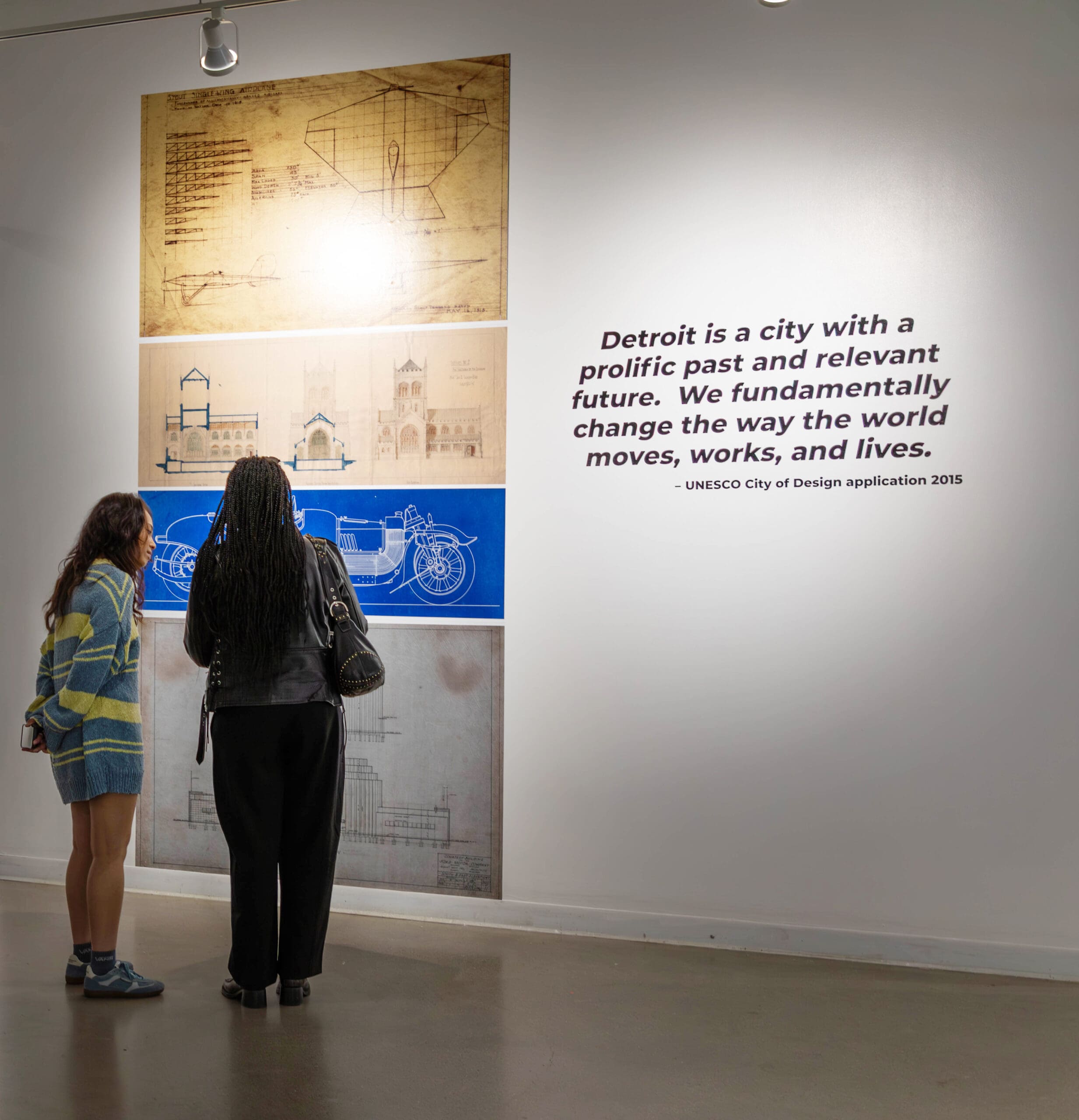 Two people standing in an art gallery, looking at an exhibition wall. On the left are four large panels displaying architectural and mechanical blueprints. On the right, a large quote on the white wall reads: "Detroit is a city with a prolific past and relevant future. We fundamentally change the way the world moves, works, and lives. – UNESCO City of Design application 2015".