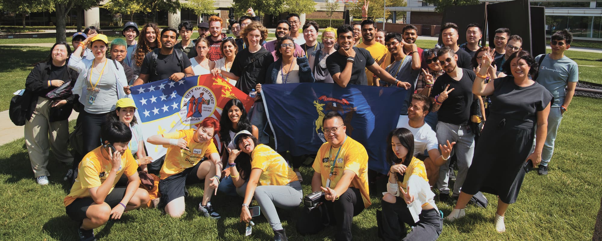 A large group of CCS international students pose outside on Ford Campus holding various flags.