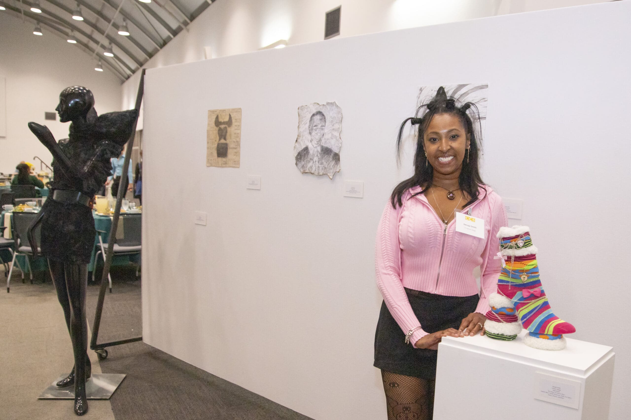 Fashion Design student Denaja Smith stands next to her colorful, striped knit footwear sculpture displayed on a white pedestal at an art exhibition.