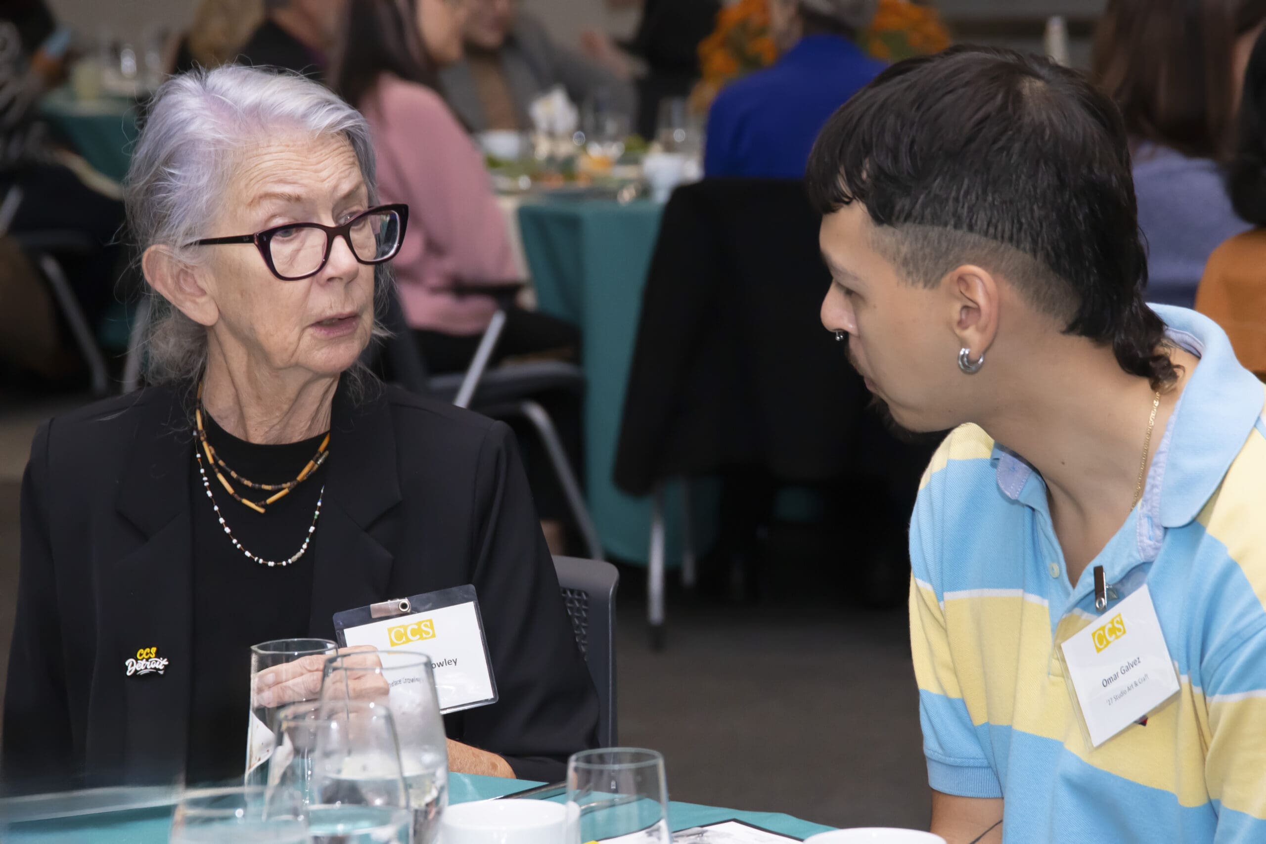 A close-up of a conversation between Candace Crowley, a woman with gray hair and glasses wearing a black blazer, and student Omar Galvez, a young person with dark hair and a yellow and blue striped shirt, sitting together at a table.