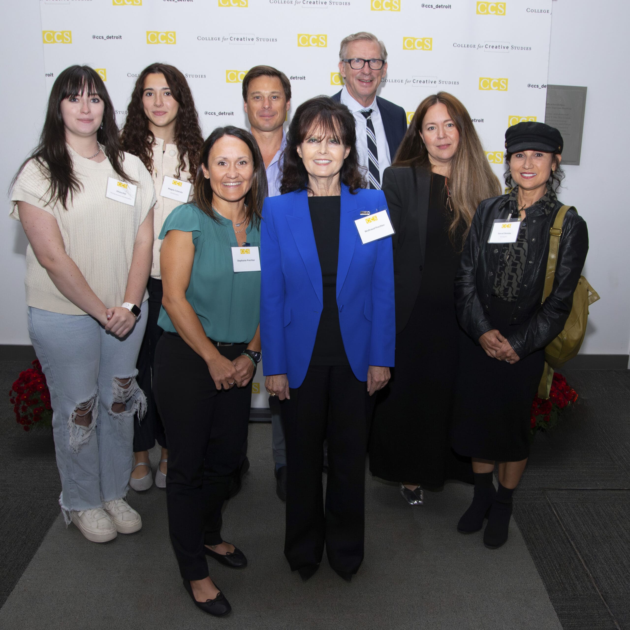 A group photo of eight people—students, alumni, faculty, and donor Waltraud “Wally” Prechter in a bright blue suit—standing together in front of a white wall branded with the CCS logo.