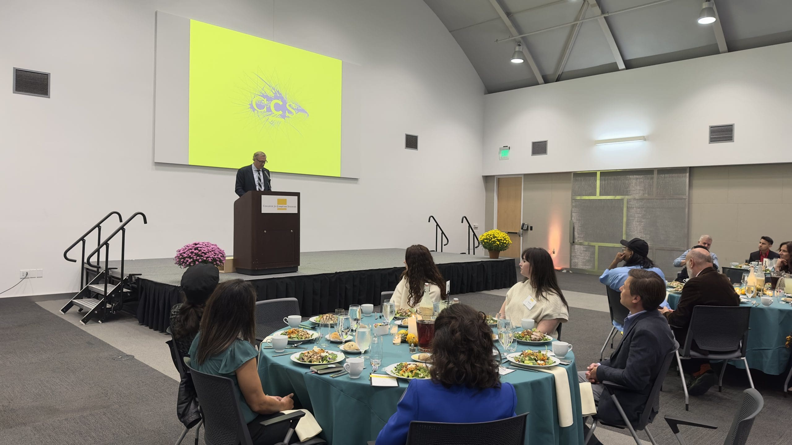 CCS President Don Tuski stands on a stage and speaks at a podium below a bright yellow screen displaying the CCS logo, addressing two teal-covered dining tables filled with attentive luncheon attendees.